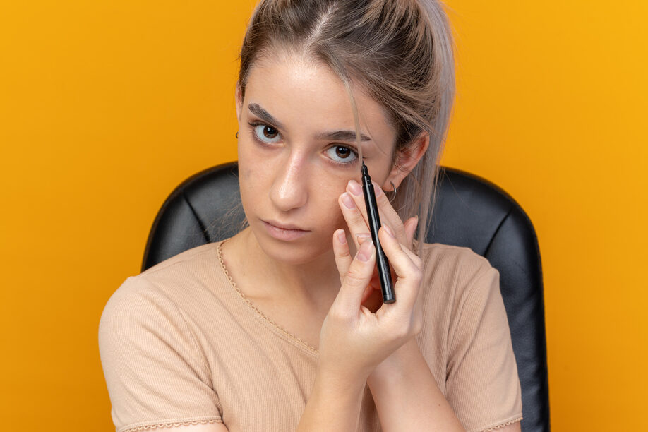 looking at camera young beautiful girl sits at table with makeup tools draw arrow with eyeliner isolated on orange background