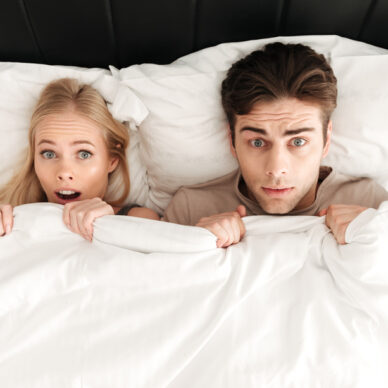 Portrait of shocked handsome couple lying in bed and cover with blanket at home