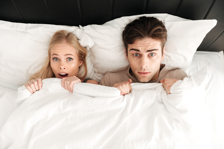 Portrait of shocked handsome couple lying in bed and cover with blanket at home