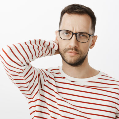 Tired and disappointed adult male employer in black glasses, touching back of head and frowning, looking focused at camera, trying concentrate on problem or question, confused over gray wall.