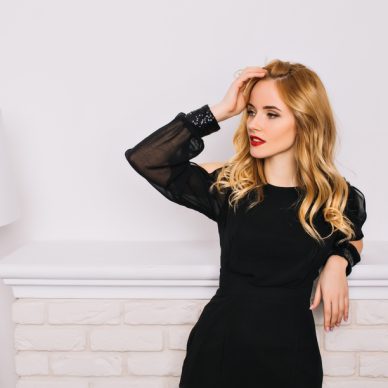 Portrait of pretty girl, young woman with blonde wavy hair sensually looking to side touching her hair. Wearing stylish black dress. White wall, fireplace, lamp on background.