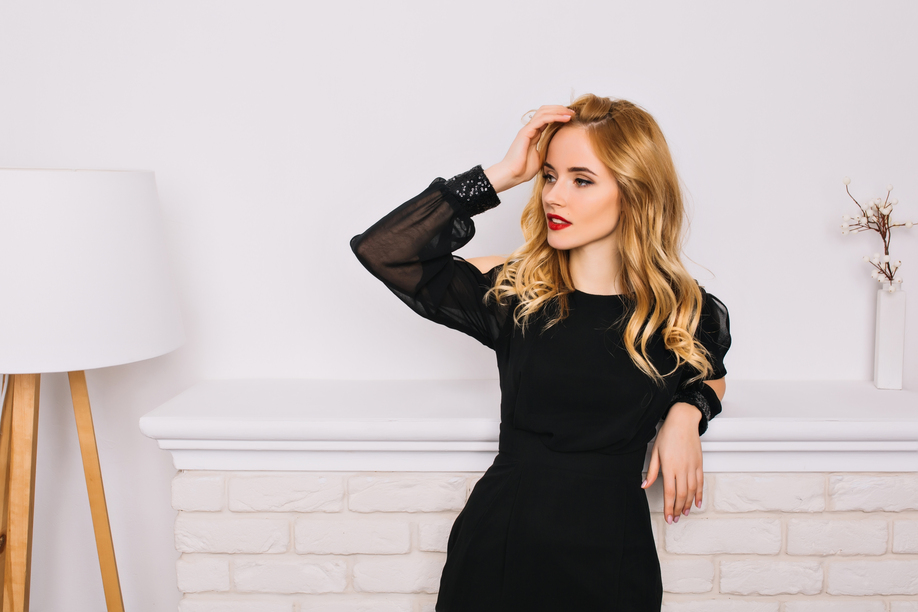 Portrait of pretty girl, young woman with blonde wavy hair sensually looking to side touching her hair. Wearing stylish black dress. White wall, fireplace, lamp on background.