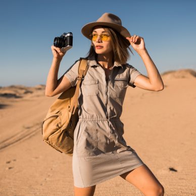 stylish young woman in khaki dress walking in desert, traveling in Africa on safari, wearing hat and backpack, taking photo on vintage camera, exploring nature, hot summer day, sunny weather