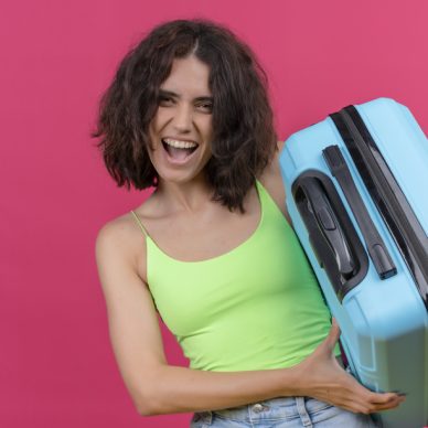 a happy lovely woman with short hair wearing green crop top showing her blue suitcase on a pink background
