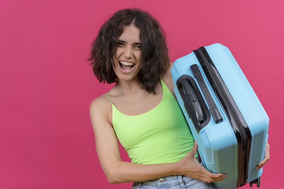 a happy lovely woman with short hair wearing green crop top showing her blue suitcase on a pink background