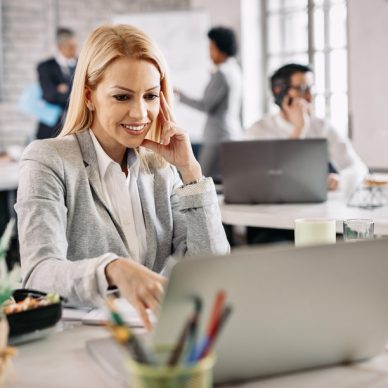 Happy mid adult businesswoman using computer and surfing on the Internet while working in the office. There are people in the background.