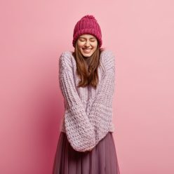 Positive Caucasian young female model keeps hands together, wears long sleeved knitted sweater, pink hat, closes eyes from pleasure, models over rosy background. People and emotions concept.