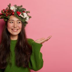 Horizontal shot of happy dark haired young woman with charming smile keeps palm raised against pink copy space, looks aside, wears beautiful holiday wreath made of spruce, demonstrates something