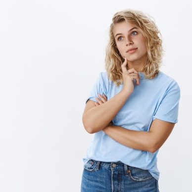 Hmm let me think. Portrait of focused attractive female friend standing in thoughtful pose looking at upper left corner as remember, recalling thought touching jawline as daydreaming over white wall.