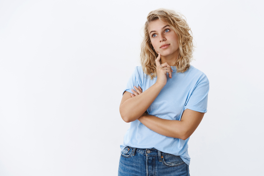 Hmm let me think. Portrait of focused attractive female friend standing in thoughtful pose looking at upper left corner as remember, recalling thought touching jawline as daydreaming over white wall.