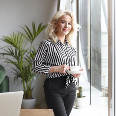 People, lifestyle, beverage, business and elderly age concept. Picture of positive successful senior woman architect having small coffee break, posing at wooden desk with laptop, hoding cup