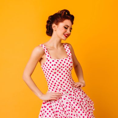 Woman in polka-dot dress posing with smile. Romantic pinup girl laughing on yellow background.