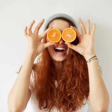 Close up studio portrait of young smiling female holding halves of oranges at her eyes. Headshot of pretty girl with loose red hair in cap and white T-shirt having fun. Film effect, selective focus