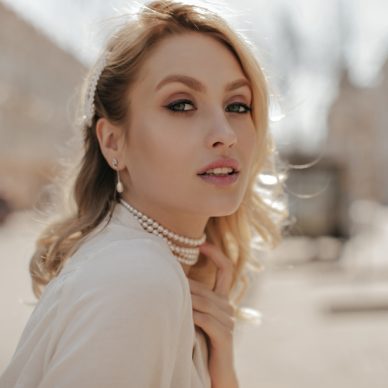 Grey-eyed beautiful woman with makeup looks into camera. Pretty girl in white blouse and pearl necklace poses outside.