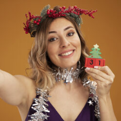 smiling young beautiful girl wearing purple dress and wreath with garland on neck holding christmas toy and camera isolated on brown background