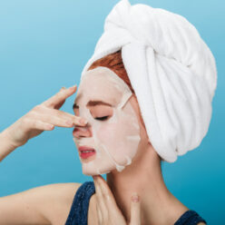 European woman with towel on head applying face mask. Studio shot of amazing girl doing spa treatment on blue background