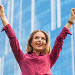 Businesswoman celebrating victory in the city centre. Pretty lady smiling with her hand raised in front of her office building. Businesswoman celebrating victory