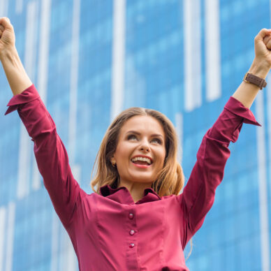 Businesswoman celebrating victory in the city centre. Pretty lady smiling with her hand raised in front of her office building.