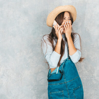 Young beautiful woman speaking on phone. Trendy shocked girl in casual summer overalls clothes and hat. Funny and surprised female posing near gray wall in studio.Closing her mouth
