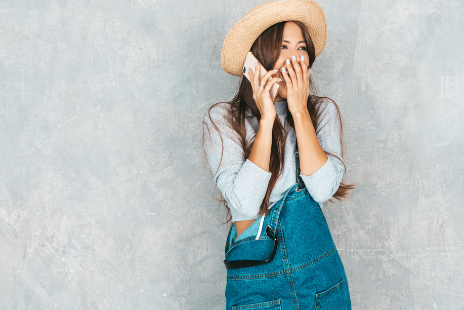 Young beautiful woman speaking on phone. Trendy shocked girl in casual summer overalls clothes and hat. Funny and surprised female posing near gray wall in studio.Closing her mouth